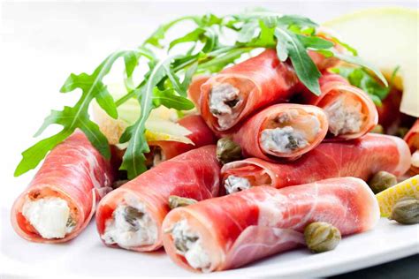 Involtini di prosciutto crudo freddi decorati con pepe rosa e verde, serviti su un piatto da portata.