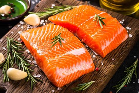 Un'immagine che mostra diversi filetti di salmone crudo, uno intero e altri già porzionati, con una varietà di erbe aromatiche e agrumi.